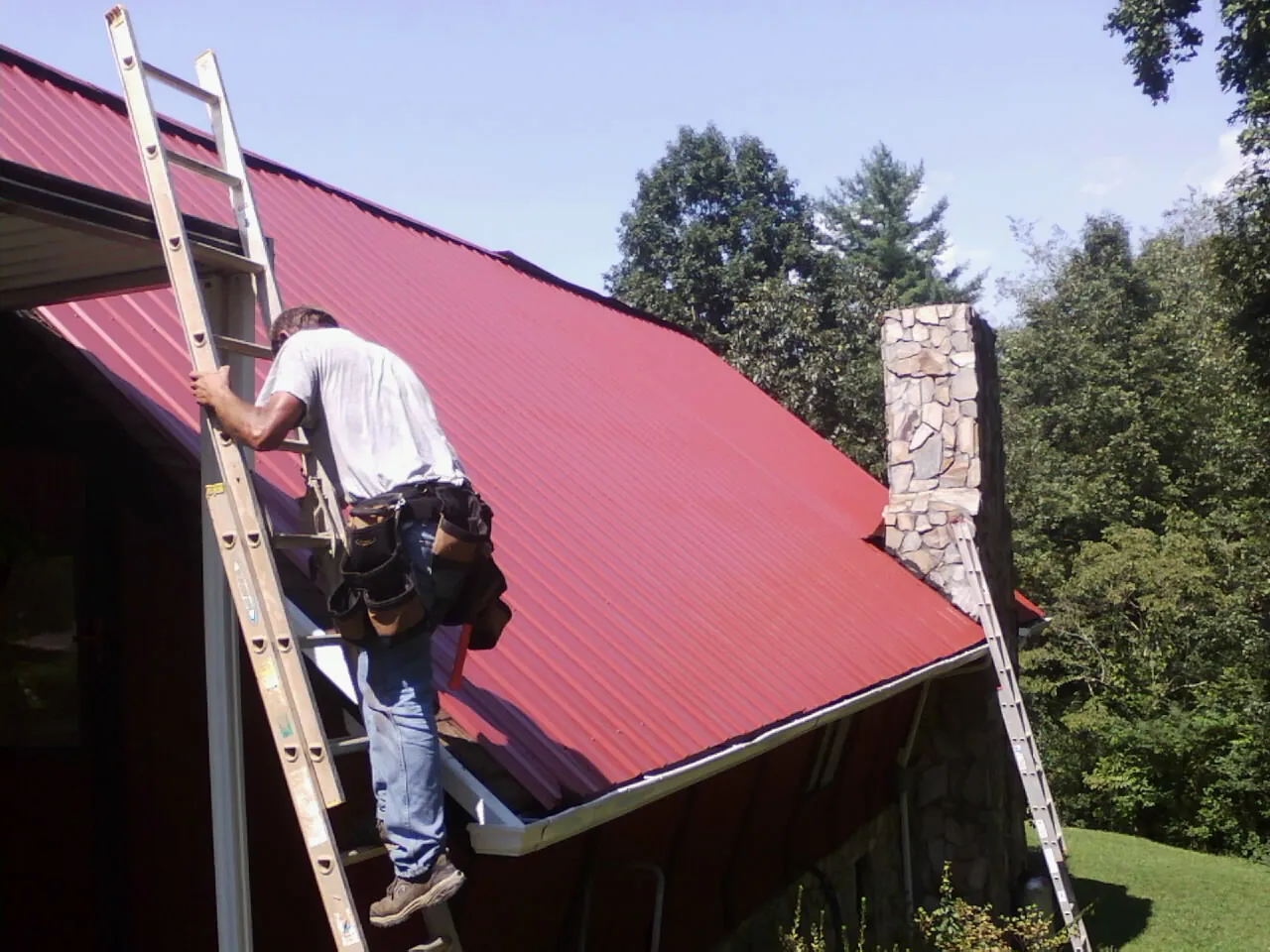Roofer on a ladder beside a red metal roof and stone chimney — Soffit & Fascia Repair by Roofing Brothers in Hillsborough